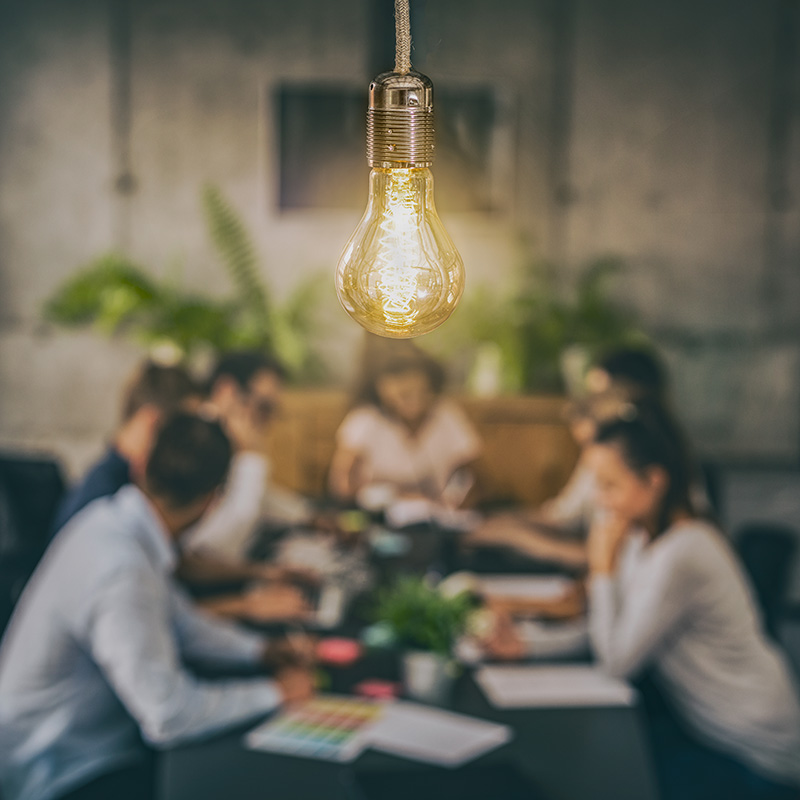 A blurred group of individuals sits at a table working while a lit lightbulb hangs front and centre
